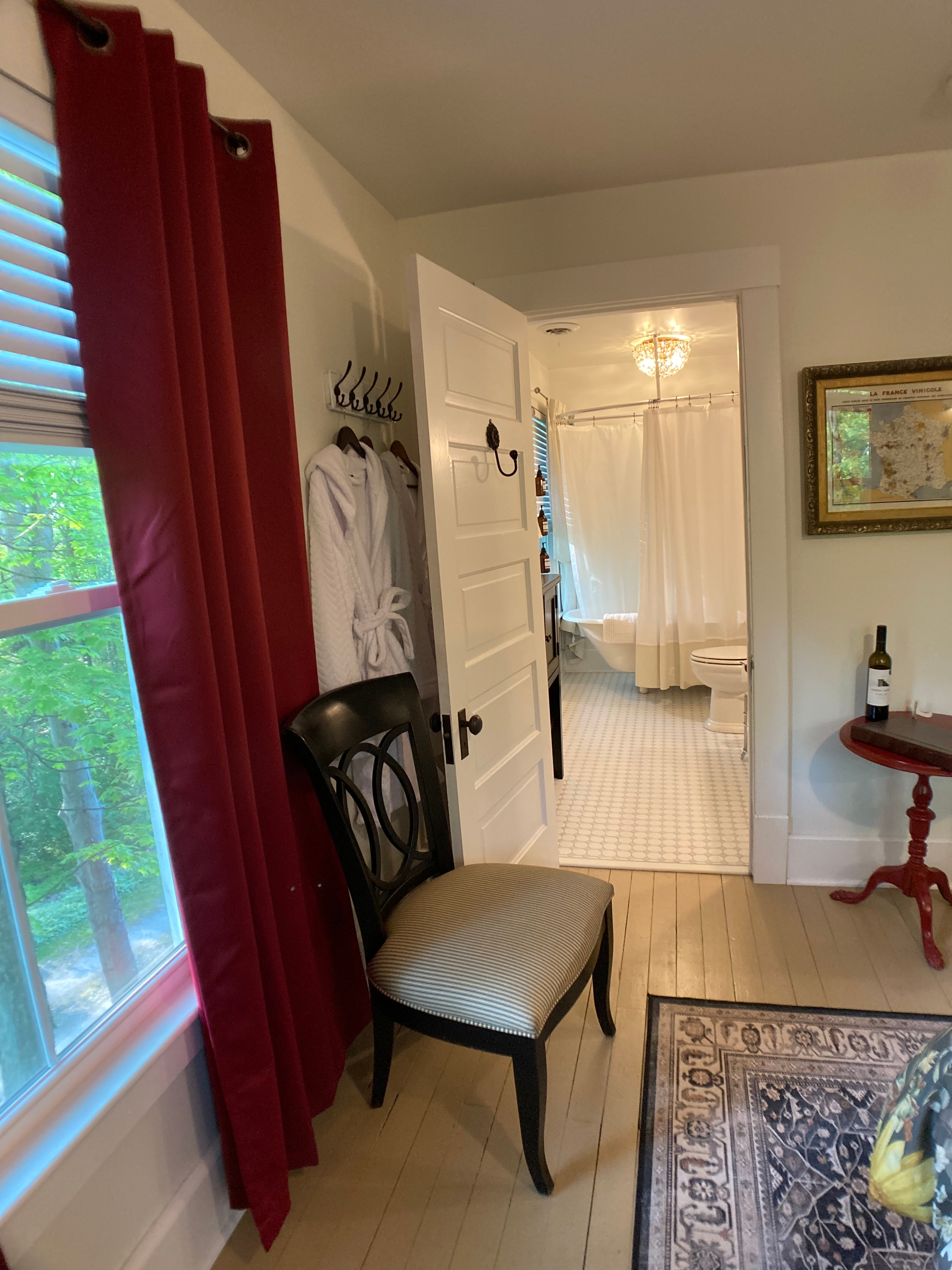 A view from the bedroom looking into the private ensuite bathroom, showing plush white robes hanging on the wall, red curtains, and a peek at the vintage clawfoot tub through the open doorway.