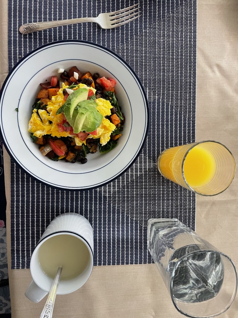 A top-down view of a fresh farm-to-table breakfast featuring a bowl of scrambled eggs topped with avocado and tomatoes, served with orange juice, coffee, and a glass of water on a blue placemat.