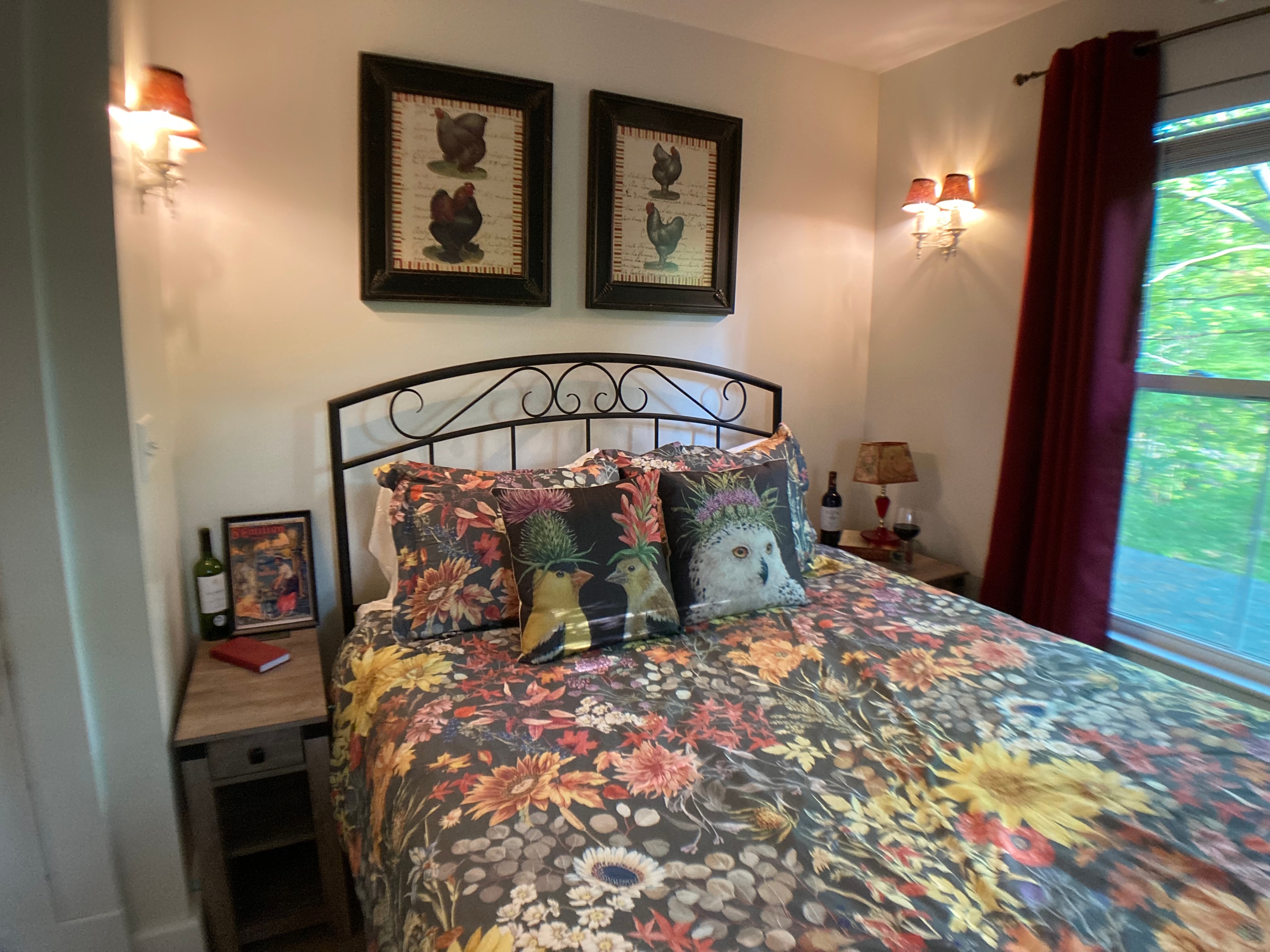 A queen bed featuring an ornate black metal headboard, floral patterned bedding with sunflowers, and whimsical throw pillows depicting chickens and an owl.