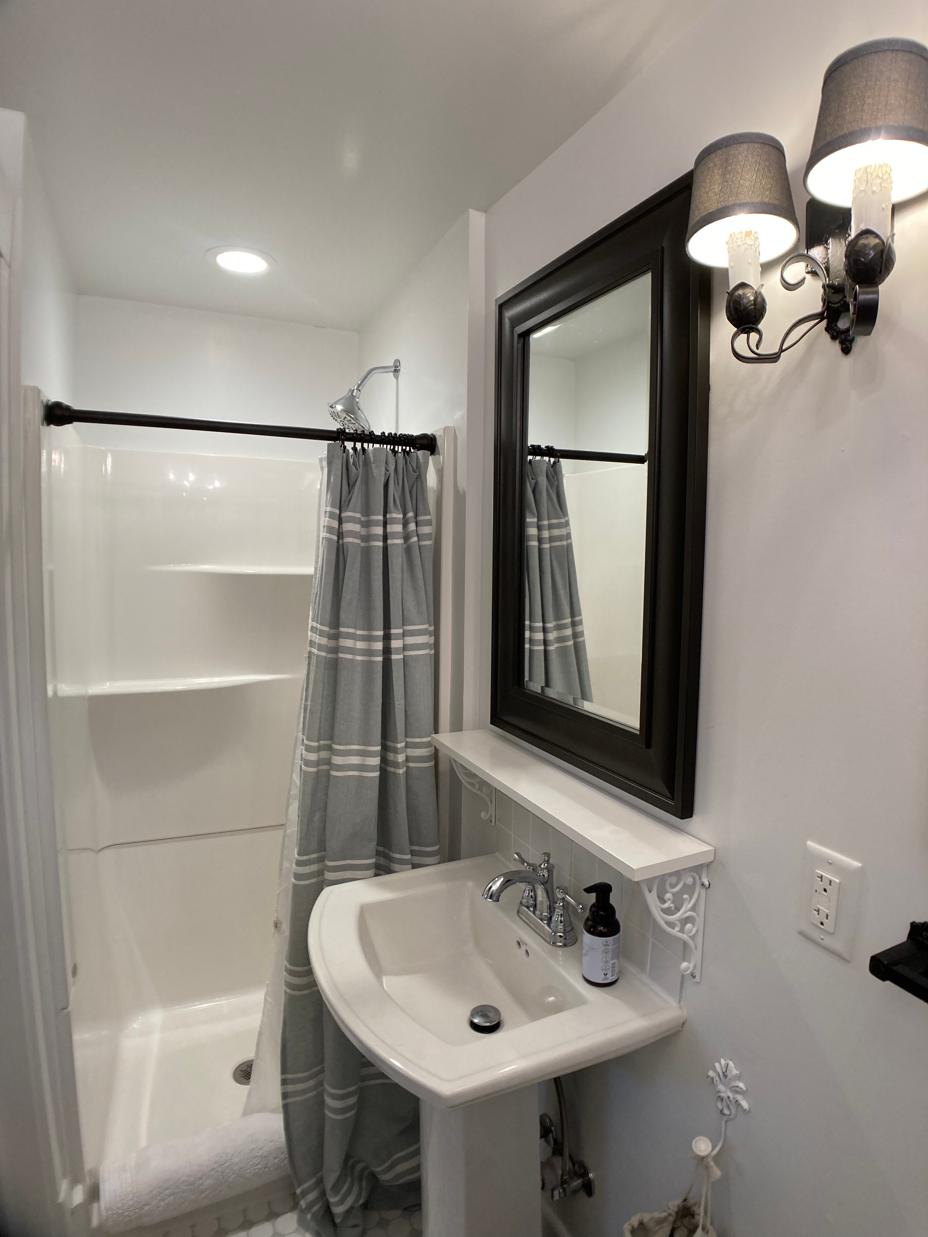 A bright ensuite bathroom with white walls and a pedestal sink. A dark-framed mirror sits above the sink, flanked by elegant black wall sconces with grey shades. A walk-in shower with a grey and white plaid curtain is visible in the corner.