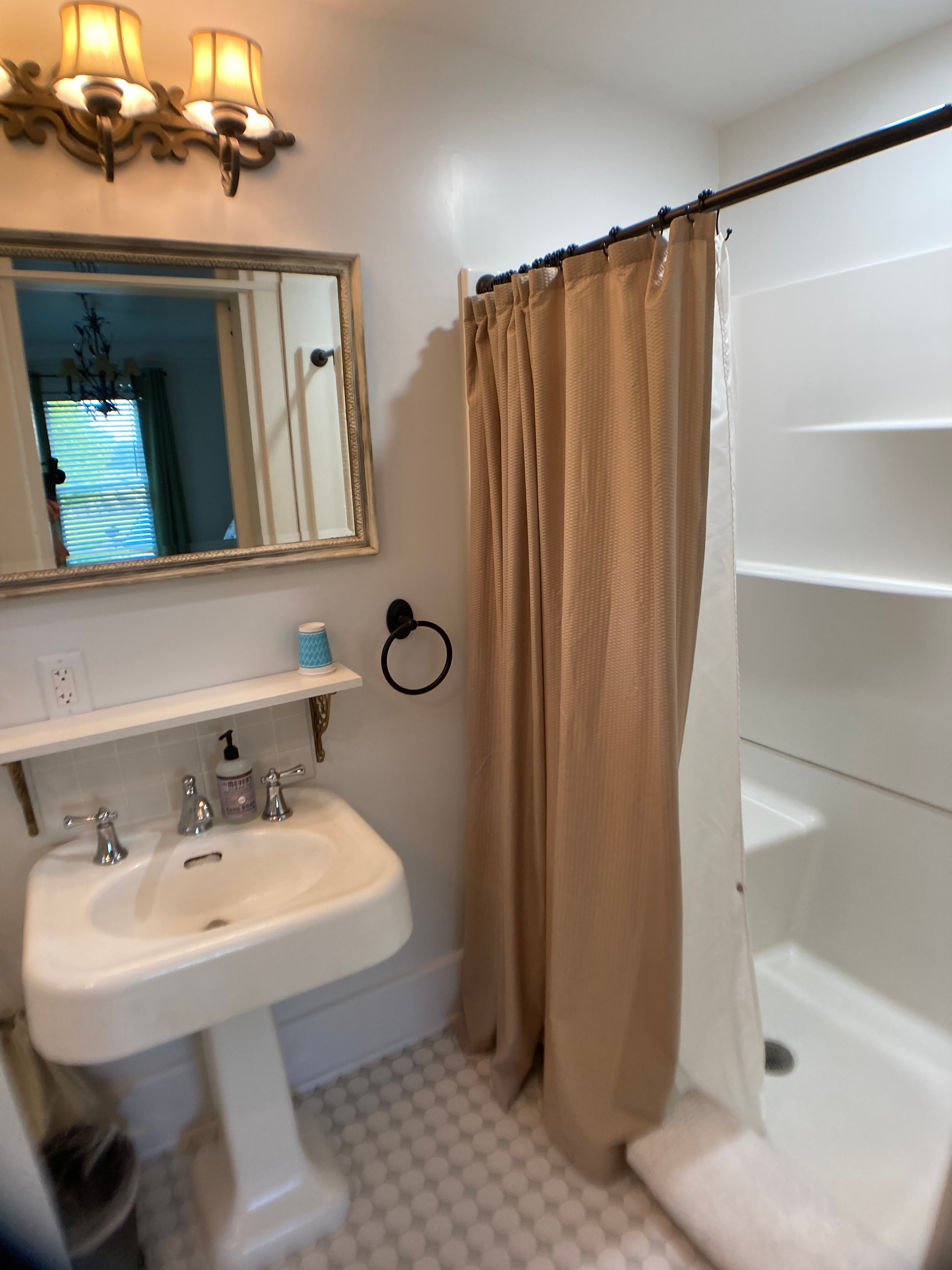 A close-up of a boutique bathroom featuring a white pedestal sink with silver fixtures, a framed rectangular mirror, and a tan shower curtain pulled across a walk-in shower.