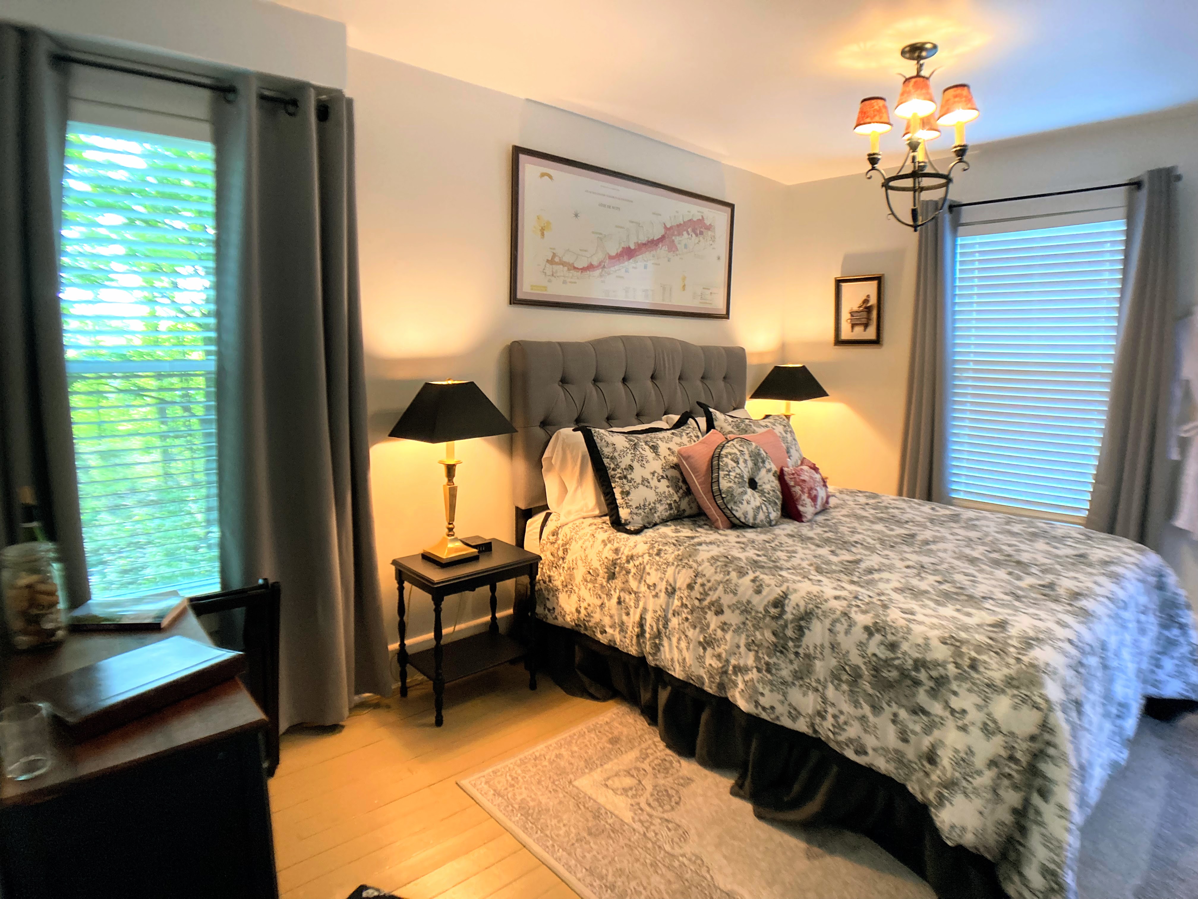 A bedroom featuring a queen bed with a grey tufted headboard, floral patterned bedding, and a framed map of a wine region. The room is illuminated by a small chandelier and matching black bedside lamps.