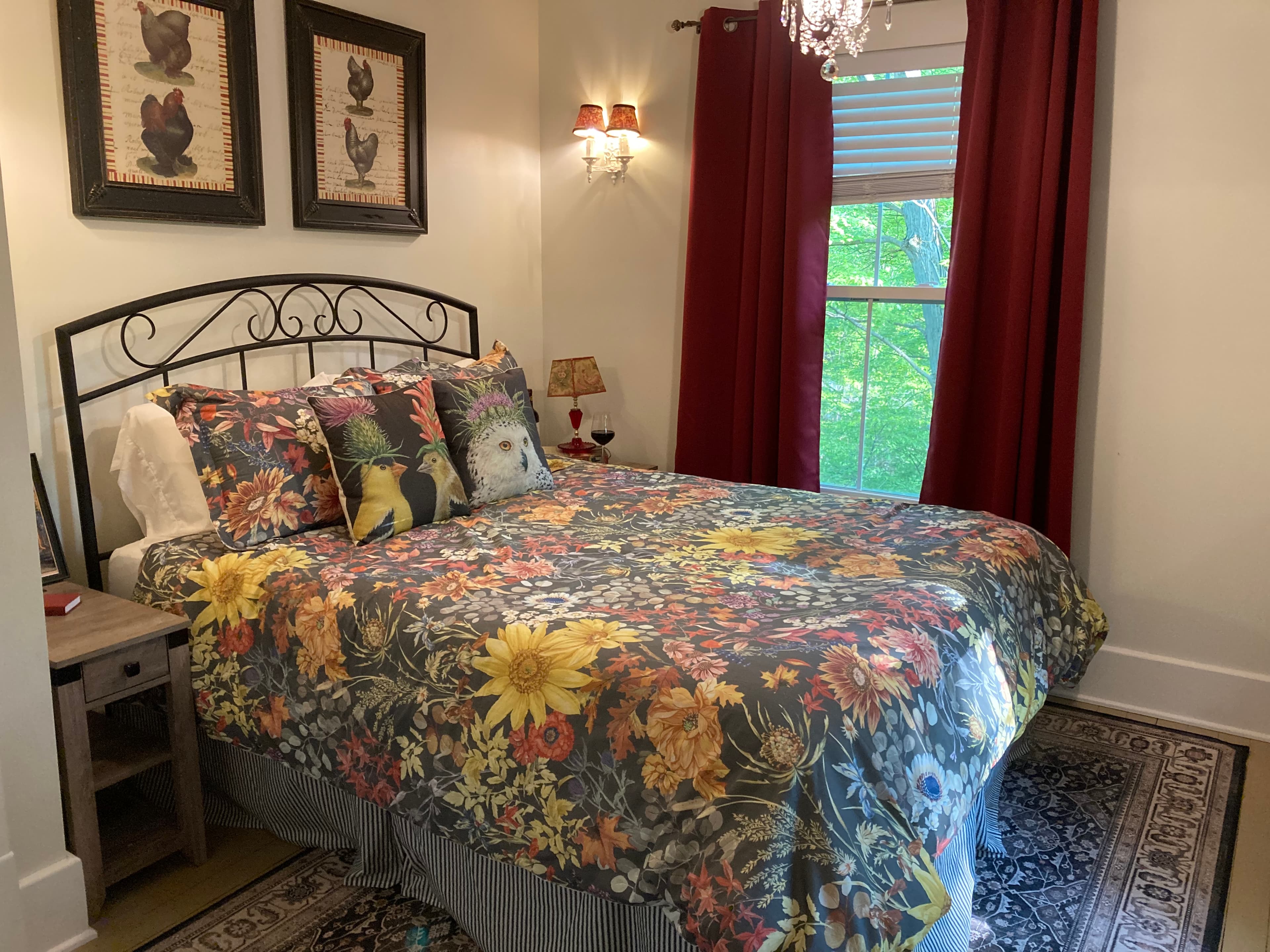 A wrought iron bed with floral bedding, framed bird artwork, red curtains, and warm lighting, creating a charming old-world atmosphere.
