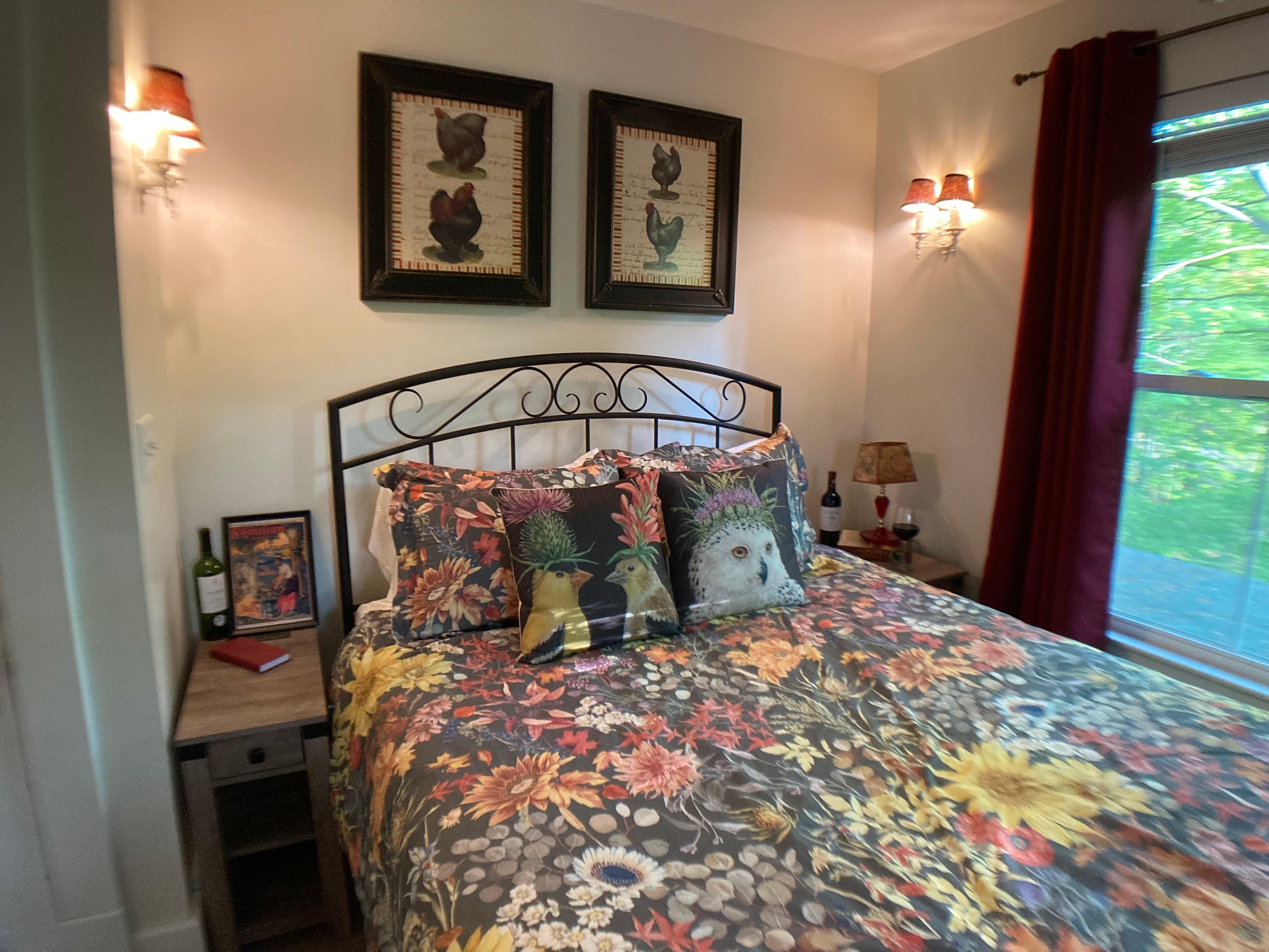 A queen bed featuring an ornate black metal headboard, floral patterned bedding with sunflowers, and whimsical throw pillows depicting chickens and an owl.