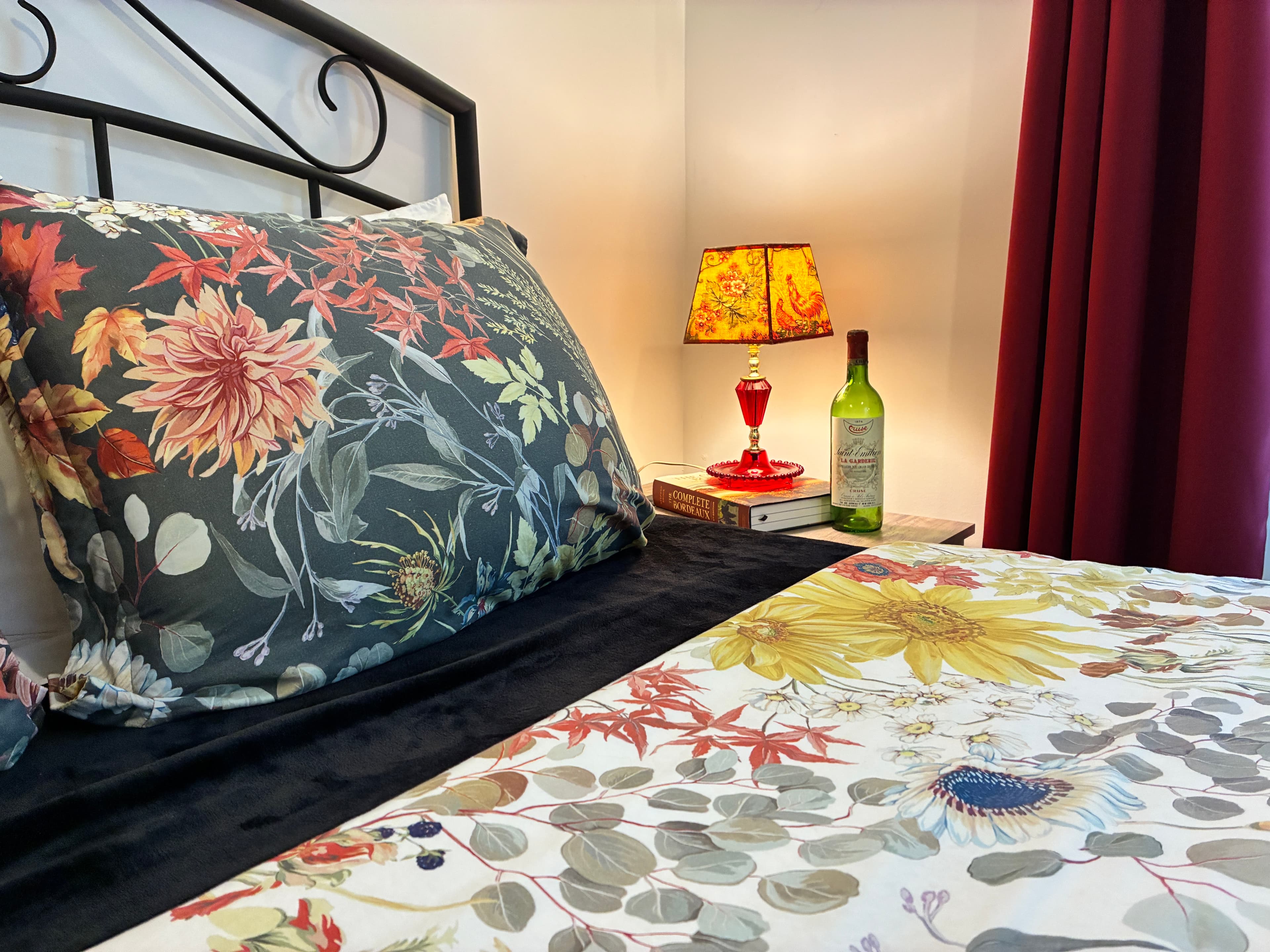 A queen bed featuring a floral pillow, a wine bottle on the nightstand, and a warm lamp glowing against deep red curtains and a wrought iron headboard.
