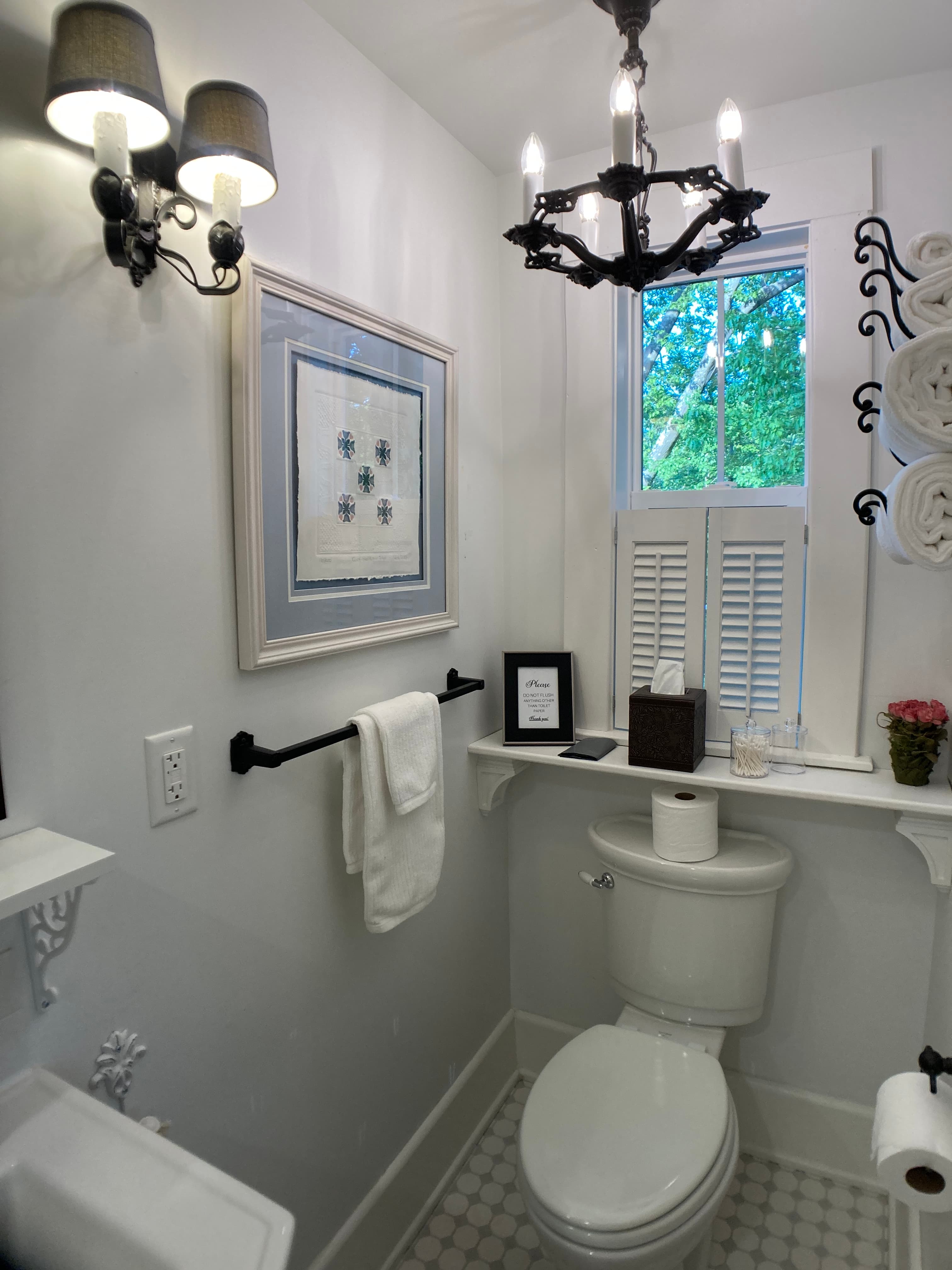 An elegant bathroom featuring white walls and a leaded glass window with white shutters. The room is accented with a black wrought iron chandelier, matching wall sconces, and a dark metal towel bar holding white towels.