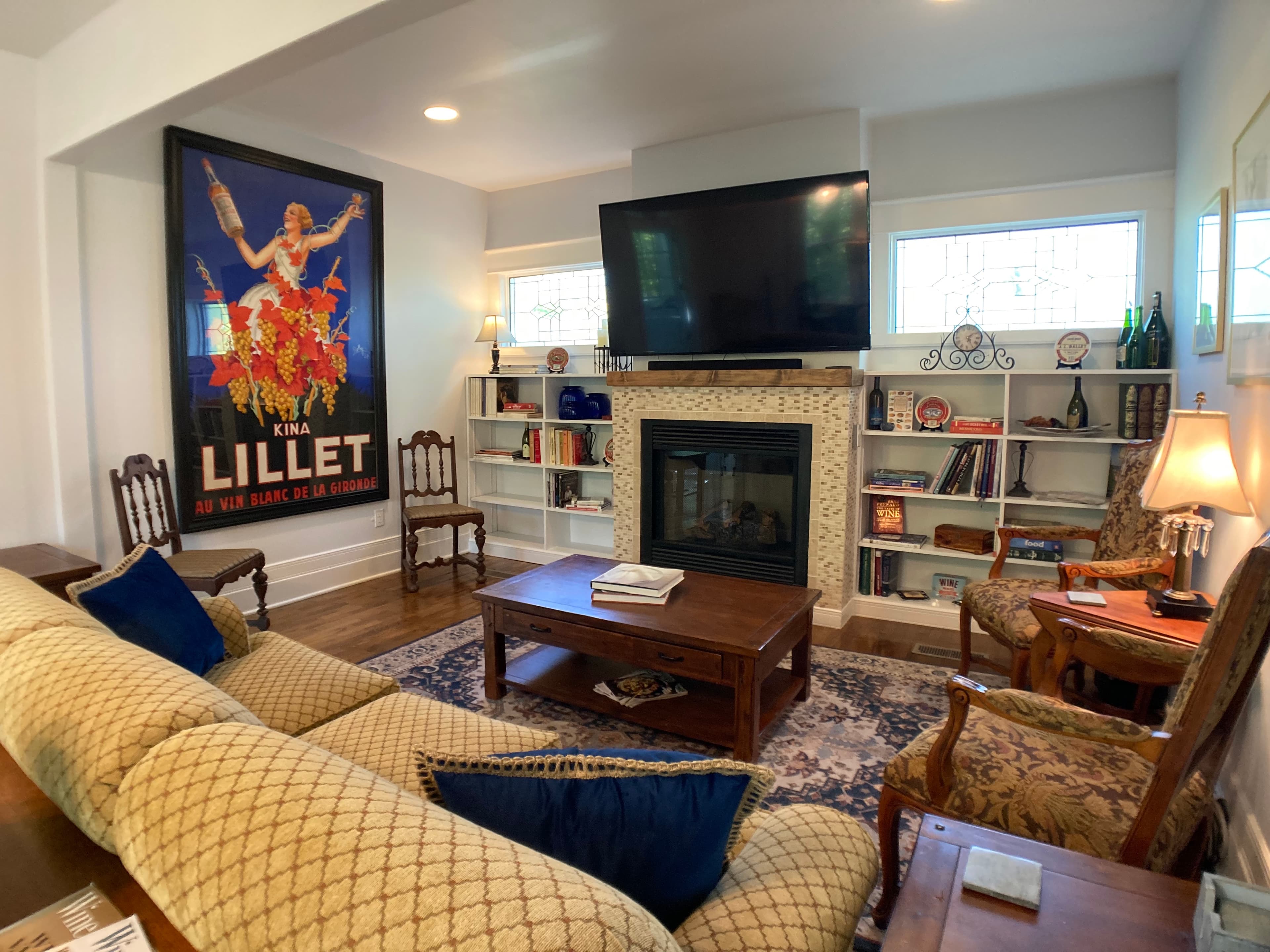 A spacious lounge featuring a large "Lillet" vintage poster, a cozy fireplace with a mounted TV, and a comfortable seating area with a tan sofa, wooden coffee table, and built-in bookshelves.