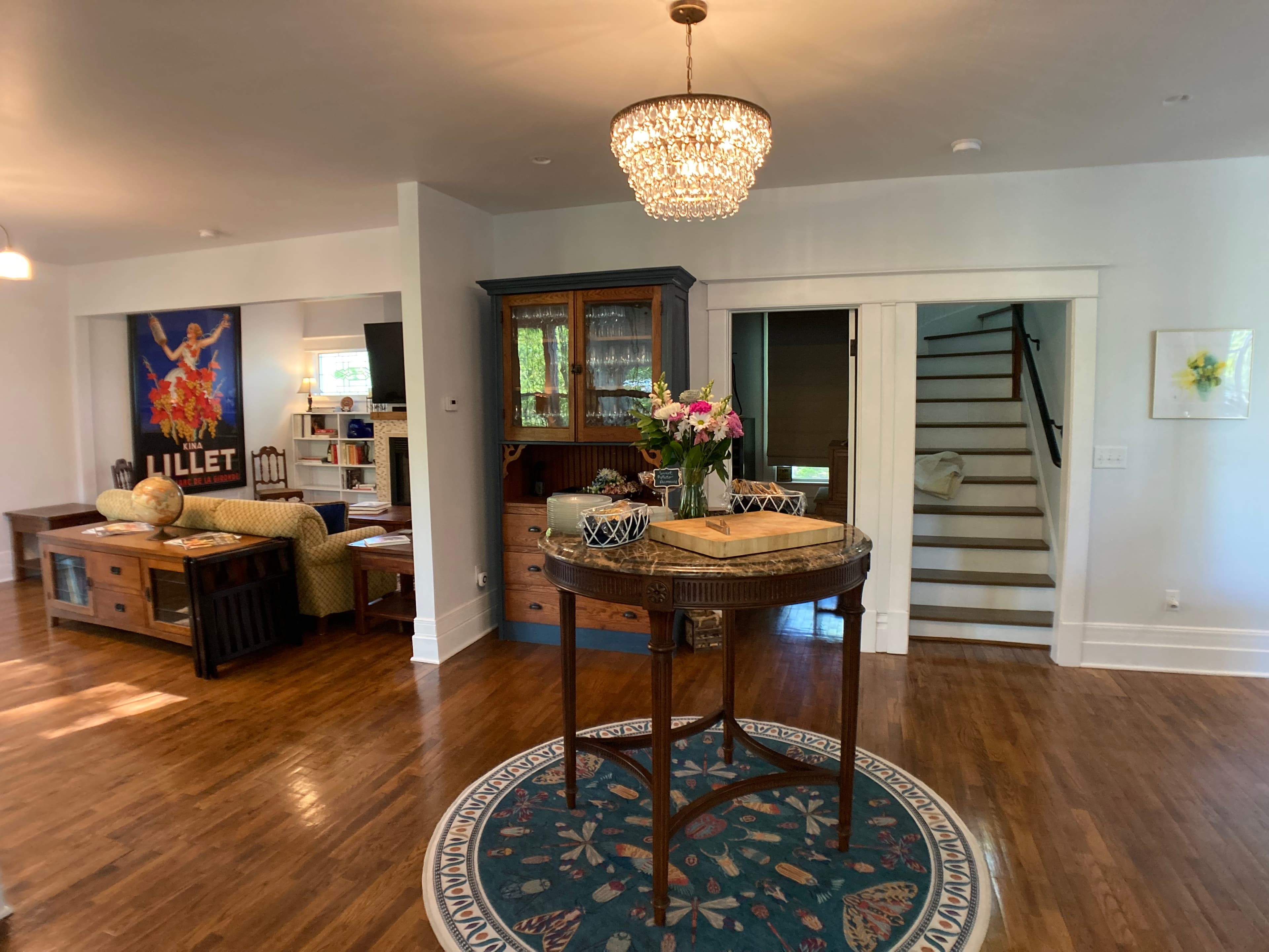 A living area featuring polished hardwood floors and a round entryway table with a floral arrangement. A crystal chandelier hangs above, with a glimpse of a staircase and a cozy lounge area with a fireplace in the background.