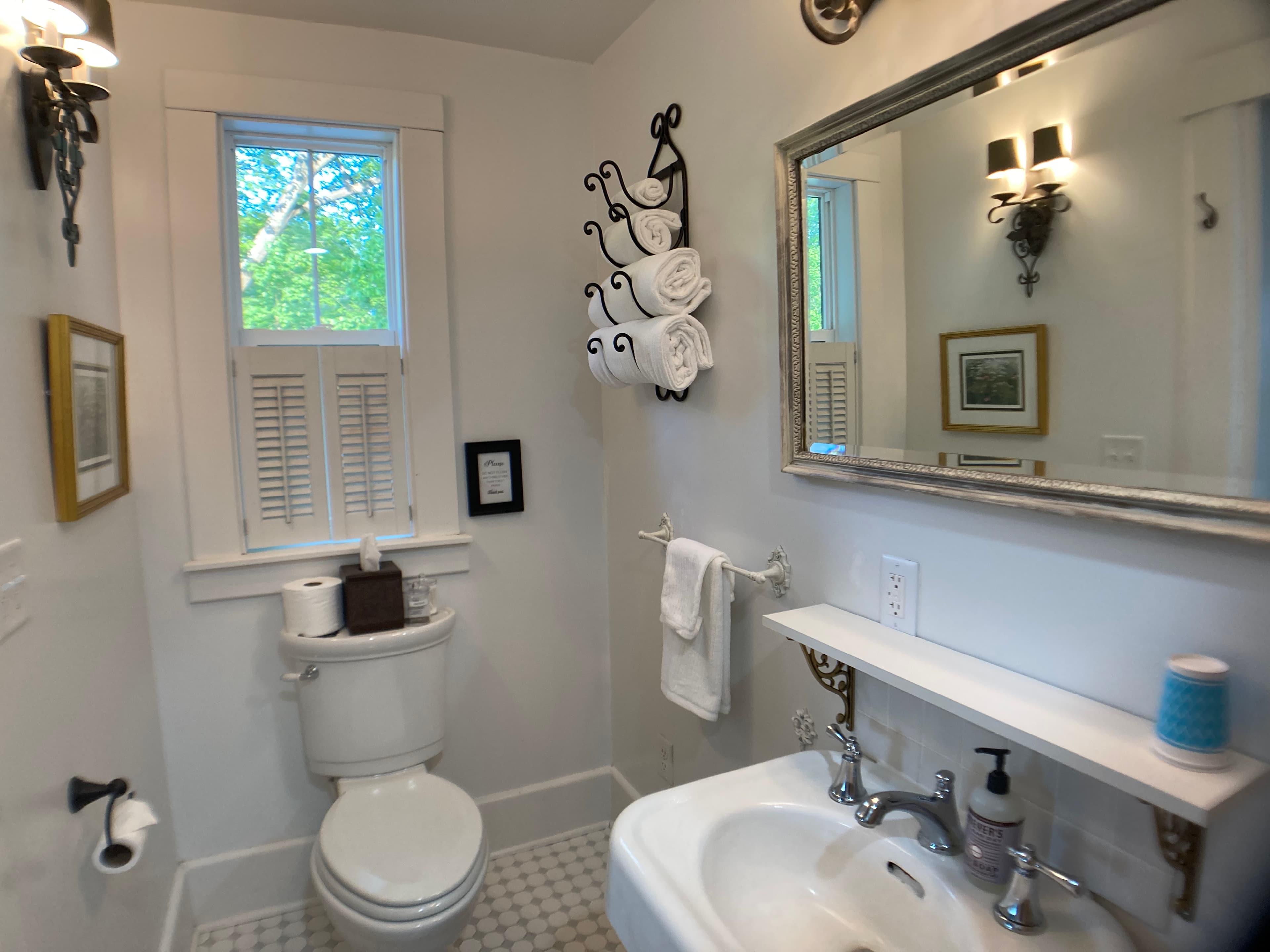 A bright, modern bathroom featuring a white pedestal sink, a silver-framed mirror, and vintage-style wall sconces. A towel rack with rolled white towels is mounted on the wall next to a window with white shutters.