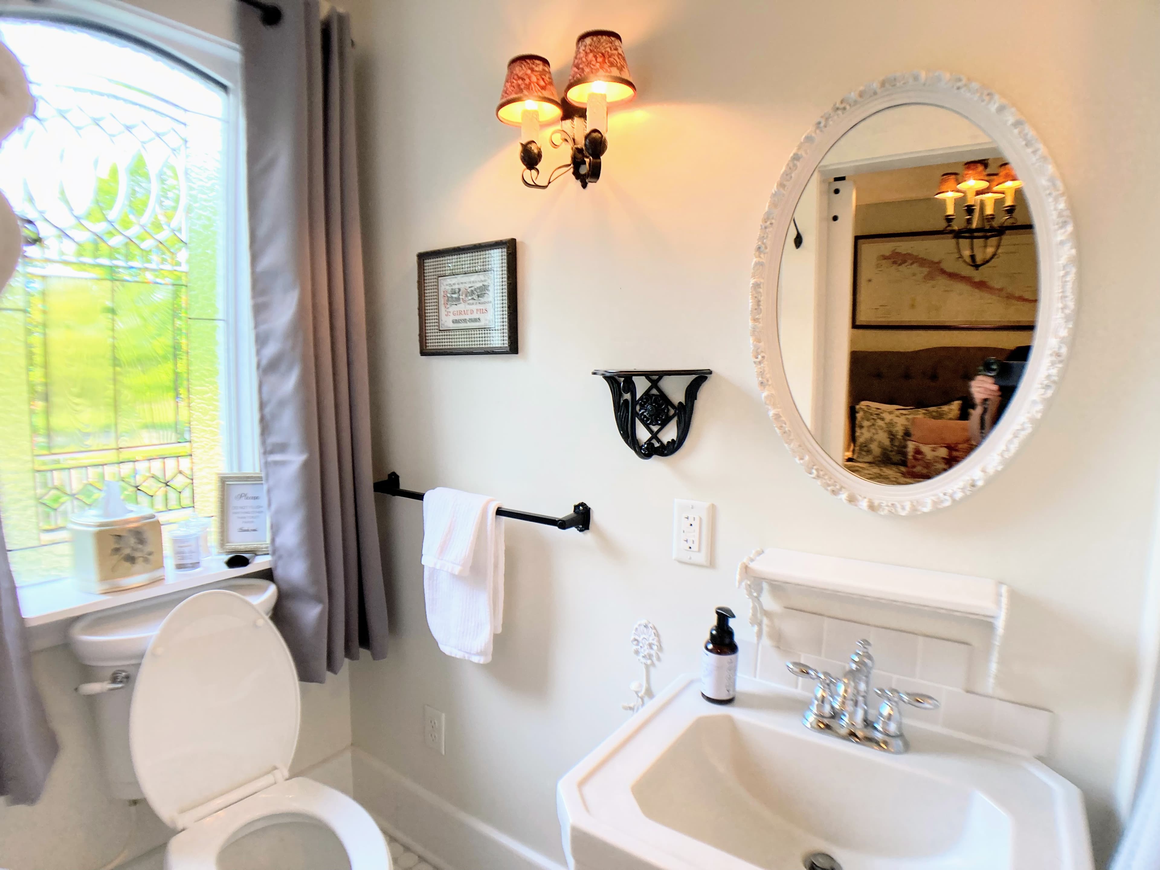 An elegant bathroom featuring a white pedestal sink and an oval-framed mirror reflecting a chandelier from the bedroom. A window with grey curtains and decorative leaded glass provides natural light, complemented by a warm three-light wall sconce.