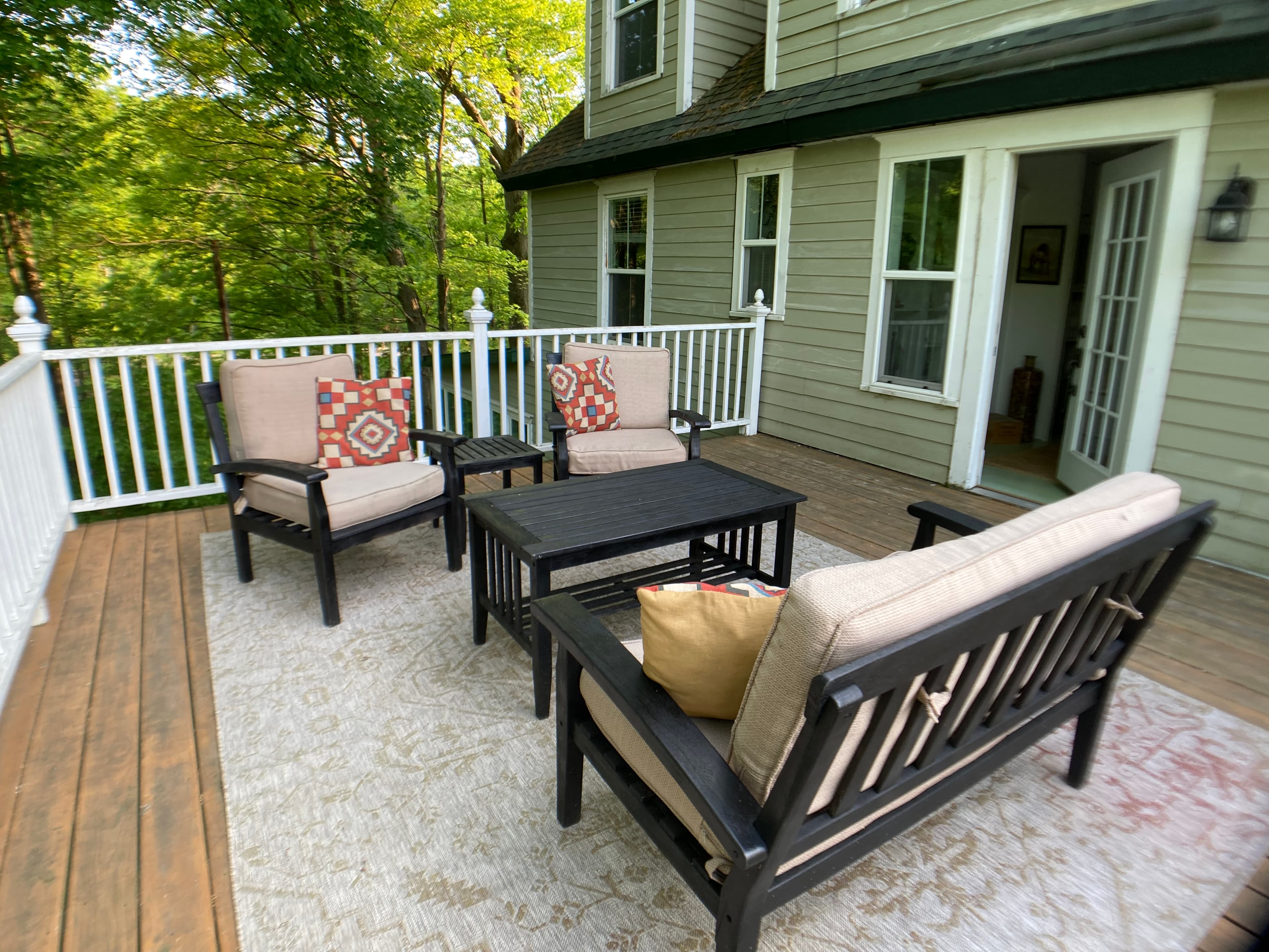 An outdoor wooden deck featuring a black patio furniture set with tan cushions and orange patterned pillows. The seating area is arranged on a light-colored rug, offering a relaxing space with views of the surrounding lush green trees.