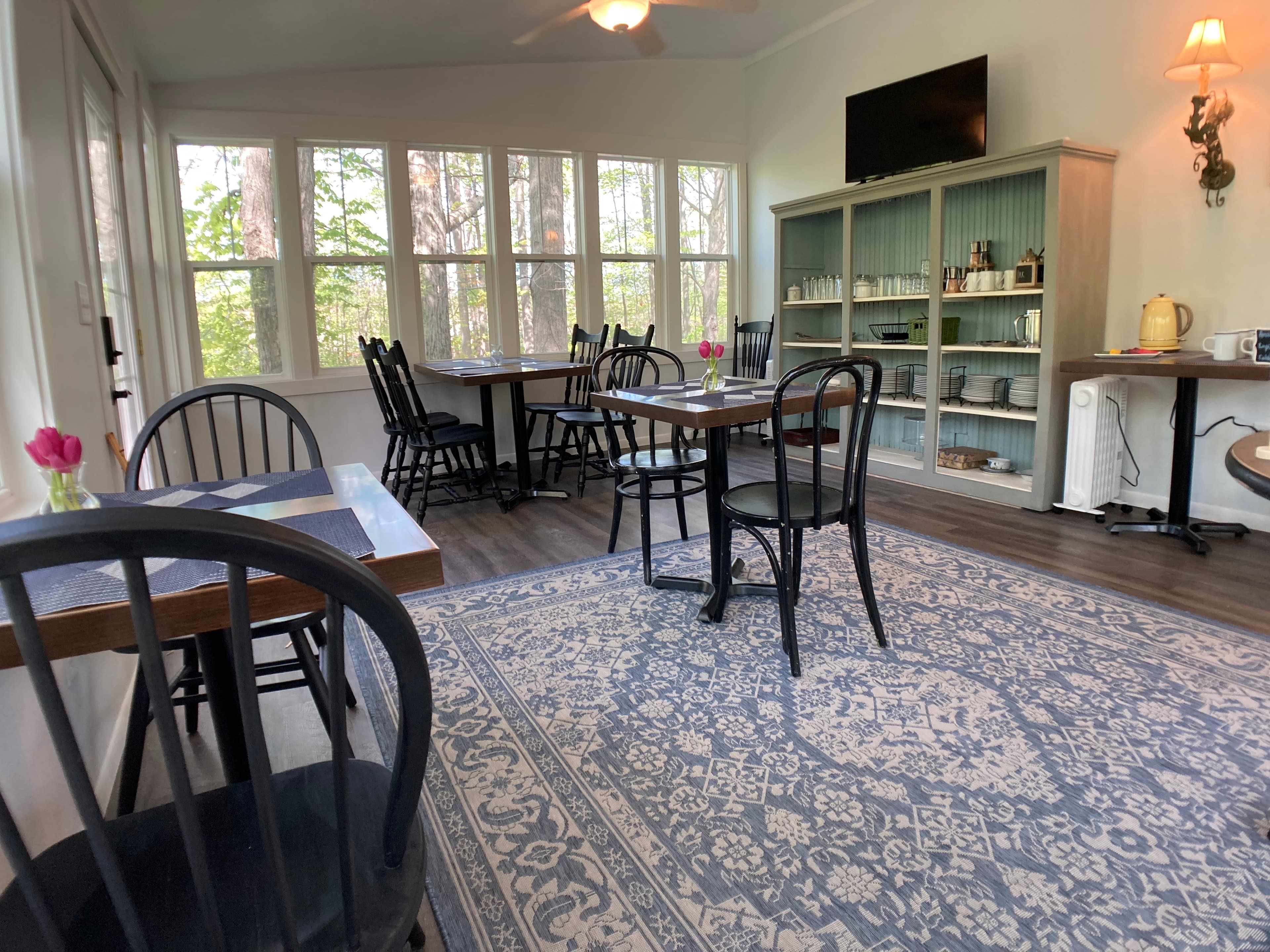 A bright dining room featuring several small wooden tables set for breakfast, a large blue and grey patterned rug, and a large window wall providing a view of the surrounding green trees.