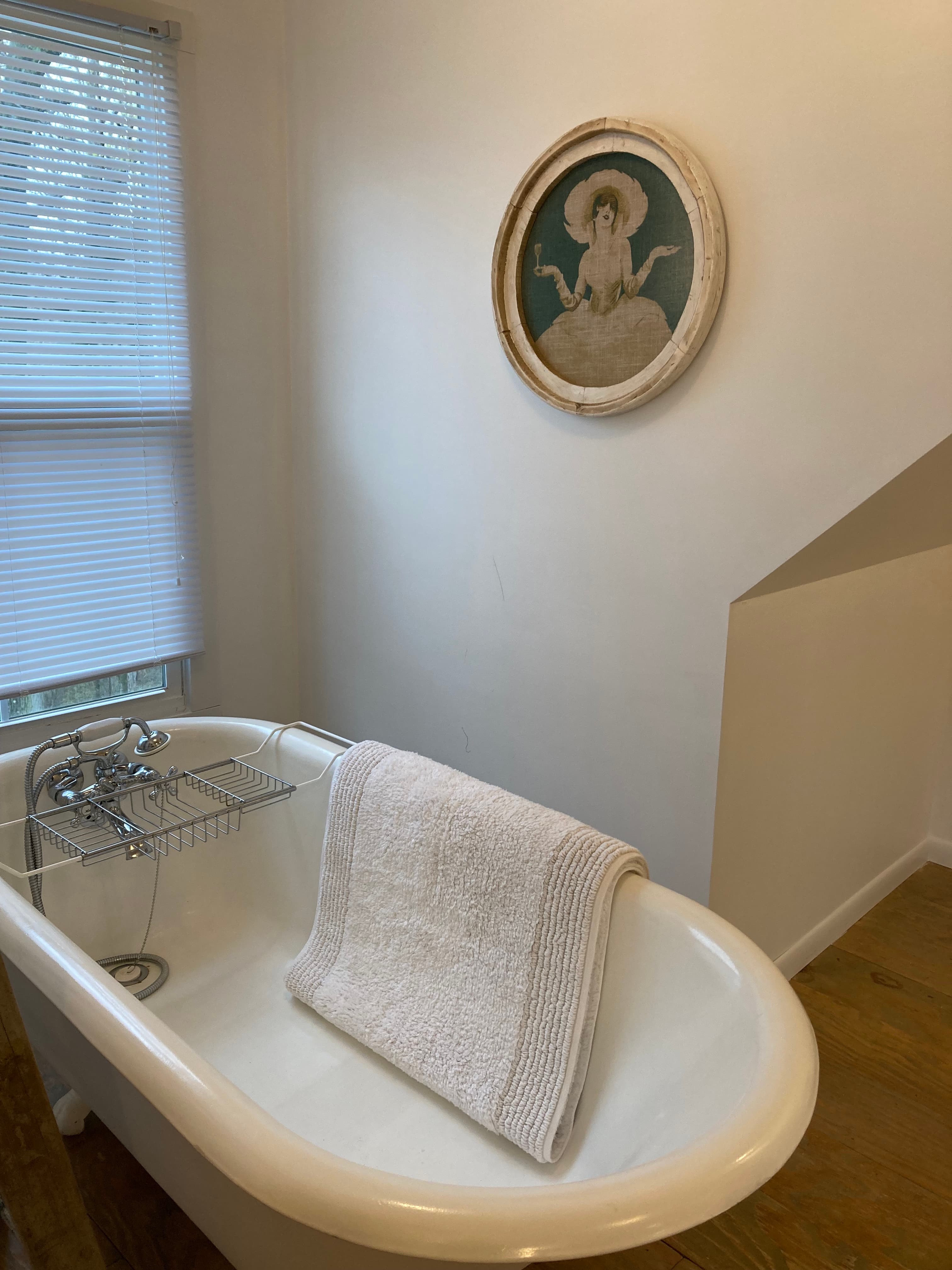A serene bathroom corner featuring a deep clawfoot soaking tub with a silver caddy and a folded white towel. A round framed artwork of a woman in vintage attire hangs on the wall above the tub.