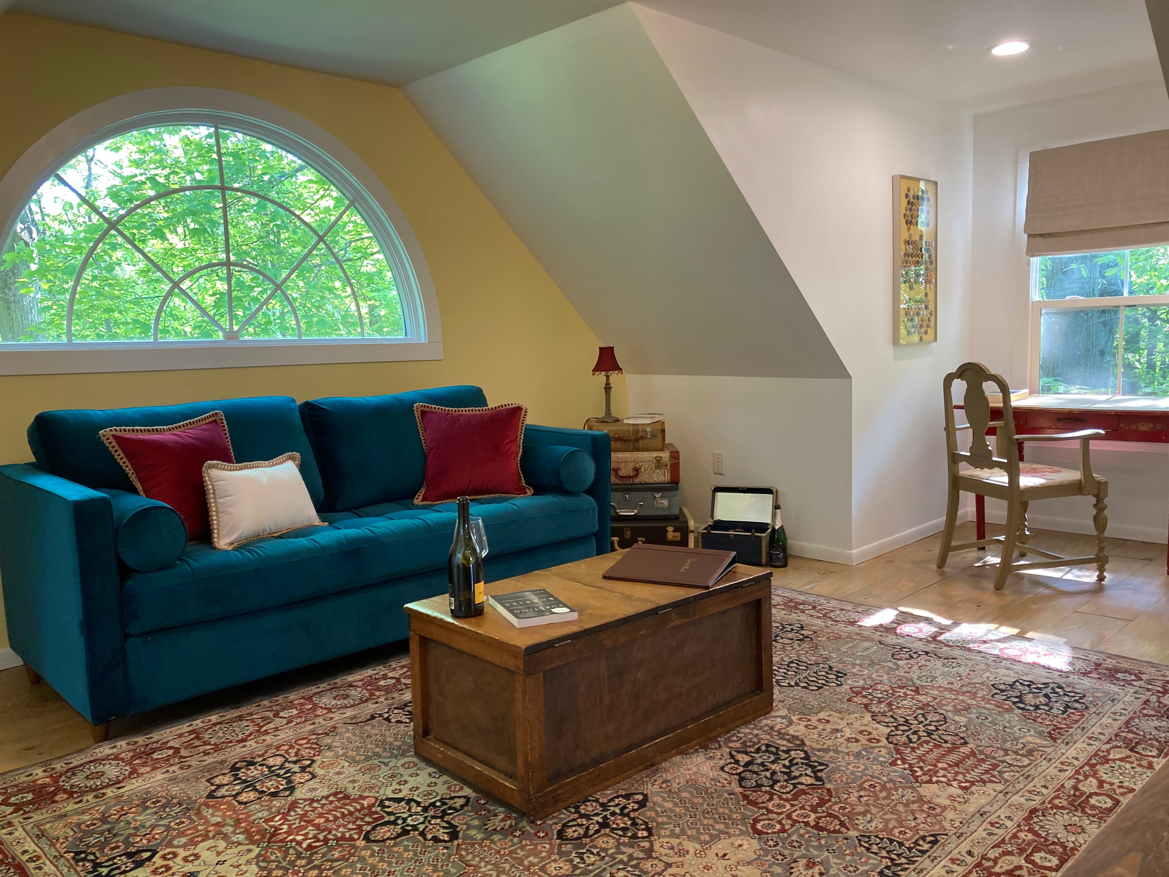 A vibrant teal velvet sofa with red and cream accent pillows sits under a large half-moon arched window. The room features sloped ceilings, a rustic wooden trunk coffee table on a patterned rug, and a small writing desk tucked into a sunlit corner.