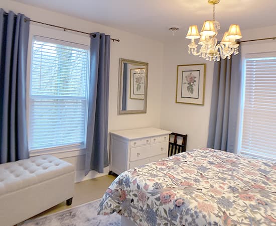 An elegant bedroom featuring a tufted grey wingback headboard, floral patterned bedding in blue and coral, and a sparkling crystal chandelier centered above the bed.