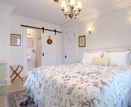 An elegant bedroom featuring a queen bed with a white and tan floral duvet, a sliding white barn door leading to an ensuite bath, and a crystal chandelier hanging from the ceiling.