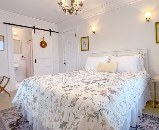 A queen bed with a floral and leaf pattern duvet, white pillows, and a sliding white barn door that leads into a bright private ensuite bathroom.