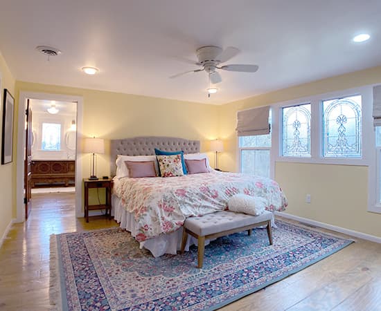 A spacious bedroom featuring a grey tufted headboard, a floral duvet with pink roses, and leaded glass windows with decorative details. The room includes a large patterned area rug, a ceiling fan, and a view into a private ensuite bathroom.