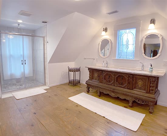 A spacious, bright bathroom featuring a large wooden antique-style double vanity with a marble top, twin oval mirrors, and a glass-enclosed walk-in shower with pebble flooring.