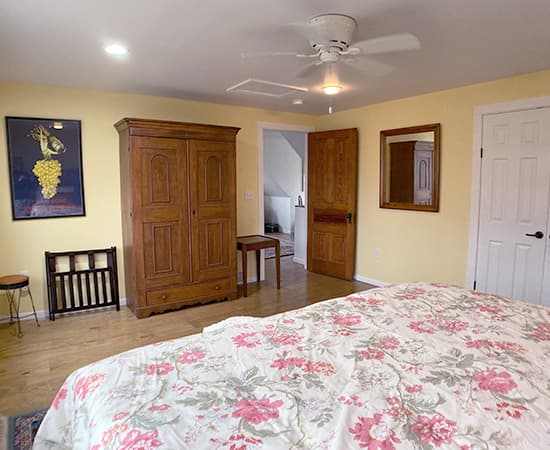 An elegant bedroom featuring a king bed with a pink floral duvet, a large wooden armoire, and a vintage wine poster on light yellow walls. Hardwood floors and a ceiling fan complete the warm, inviting space.