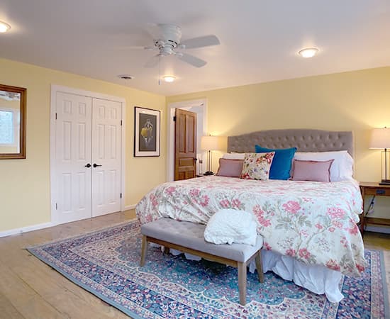 A spacious bedroom featuring a king bed with a pink floral duvet, a tufted grey headboard, and light yellow walls. A large patterned area rug sits on hardwood floors, with a large wooden armoire visible in the corner.