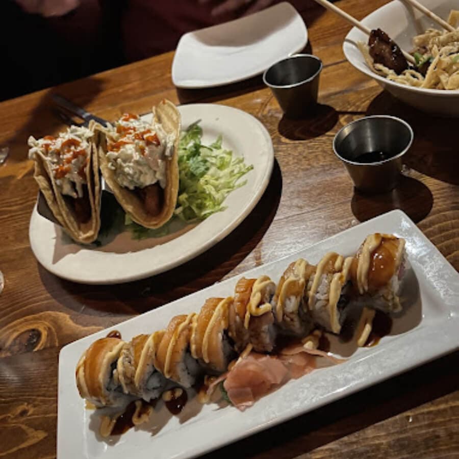 A plate featuring sushi rolls drizzled with sauce and two tacos filled with toppings, set on a wooden table.
