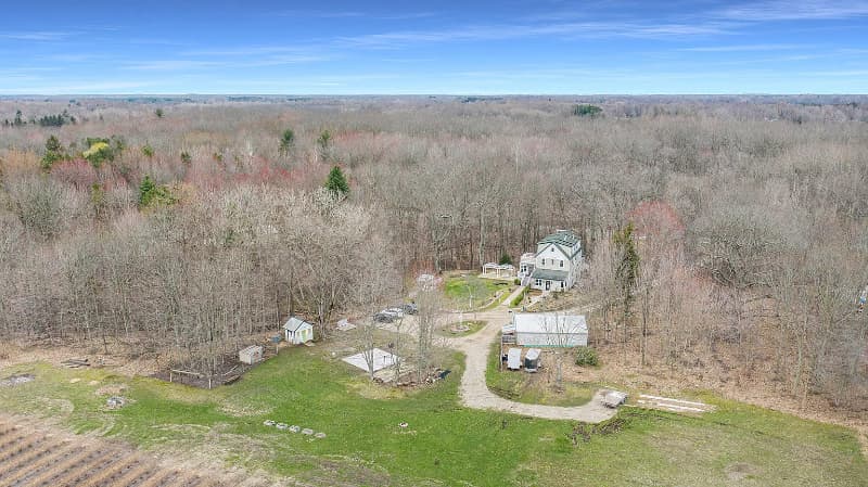 Aerial view of a rural property surrounded by dense forest, featuring a house and several outbuildings.