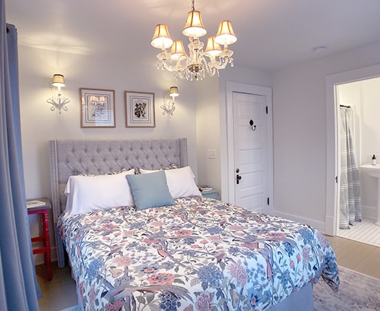 An elegant bedroom featuring a tufted grey wingback headboard, floral patterned bedding in blue and coral, and a sparkling crystal chandelier centered above the bed.