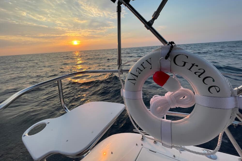 A lifebuoy inscribed with "Mary Grace" rests on a boat with a sunset over the ocean in the background.