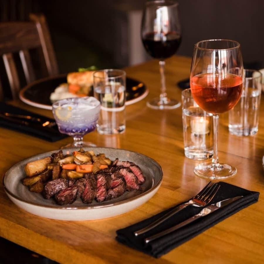 A plate of sliced steak with roasted potatoes on a wooden table, accompanied by various drinks and tableware.