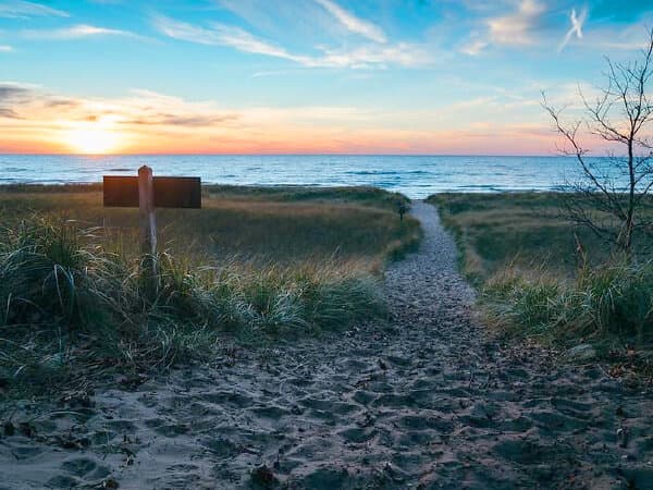 Pathway through grassy dunes leading to a tranquil beach at sunset.