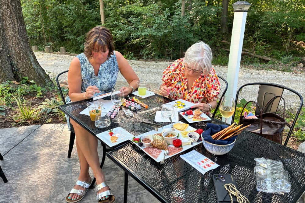 Two women enjoy a meal and drinks at an outdoor table surrounded by greenery.