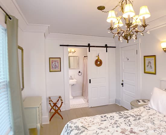 A bright boutique guest room featuring a white sliding barn door that opens to a private ensuite bathroom. The room is decorated with a floral duvet, a gold chandelier, and soft green curtains.