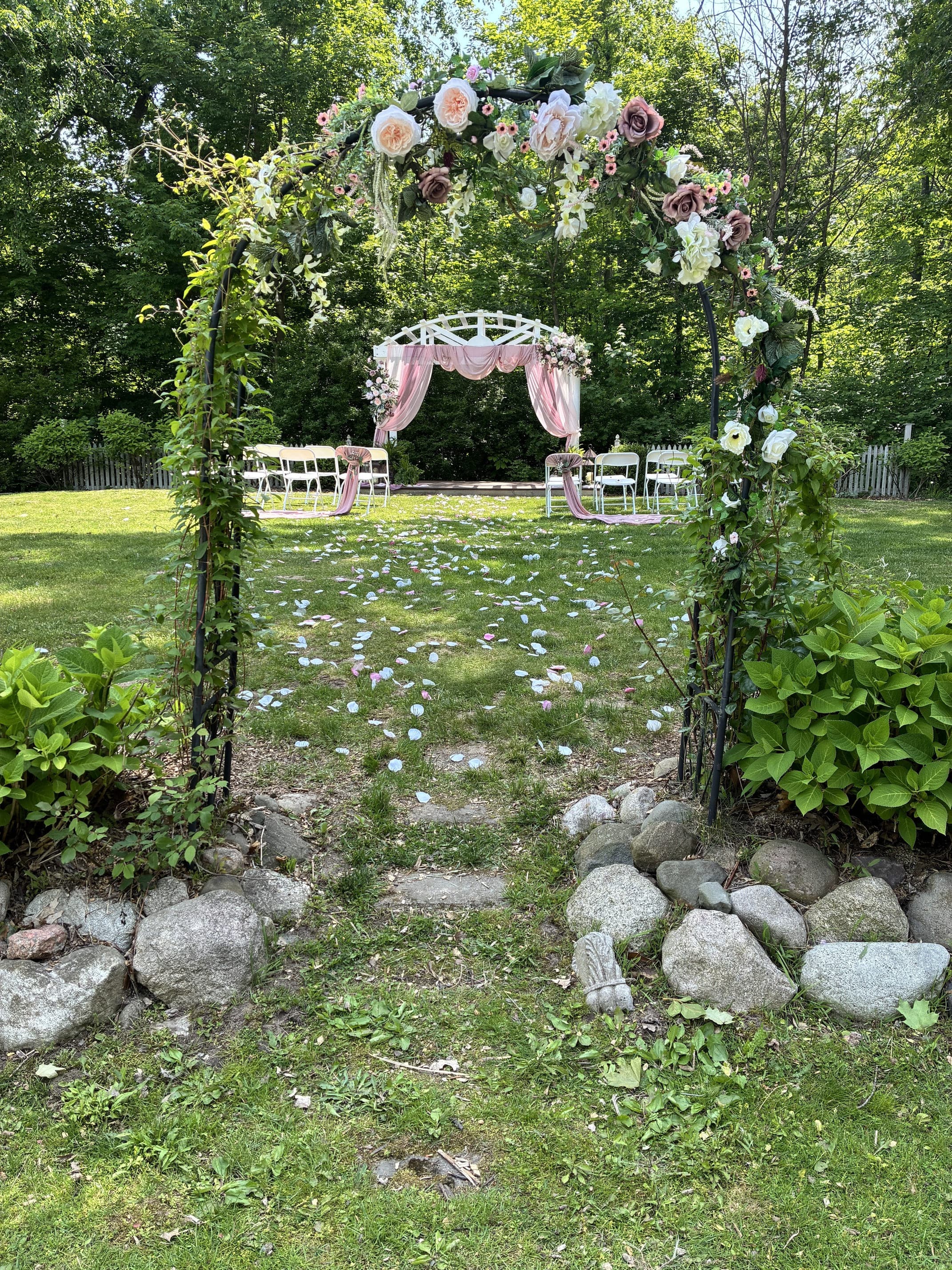 A flower-adorned archway leads to a decorated wedding altar surrounded by white chairs and scattered petals on a grassy lawn.
