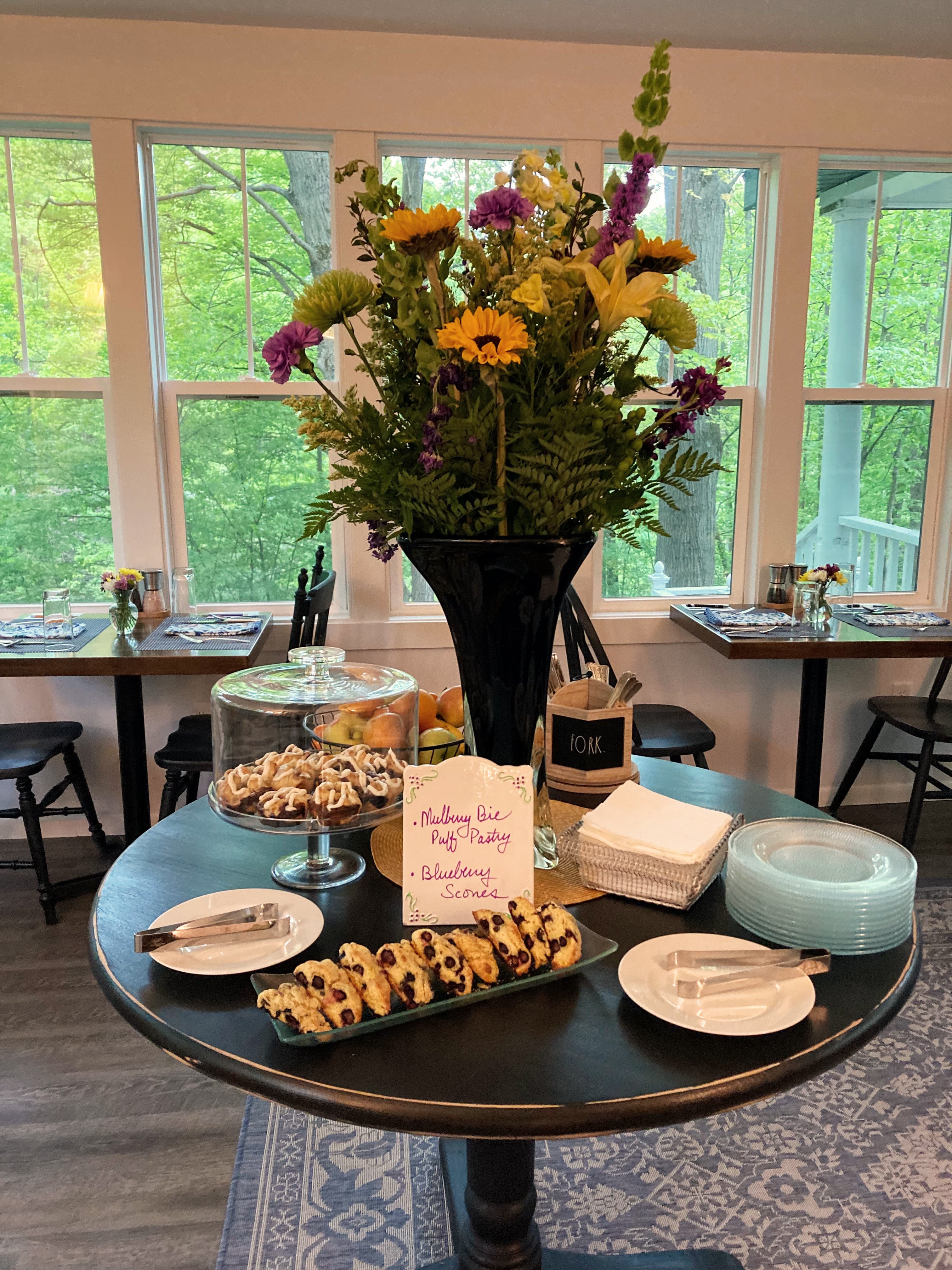 A round wooden table set for breakfast with a large floral centerpiece of sunflowers and greenery. The display includes fresh scones under a glass dome and small pastries on a tray, with views of green trees through large windows in the background.