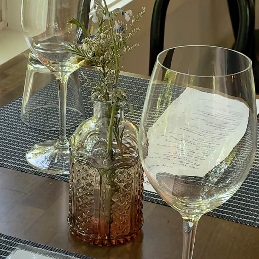 A small glass bottle with a flower arrangement sits on a set dining table beside wine glasses and a menu.