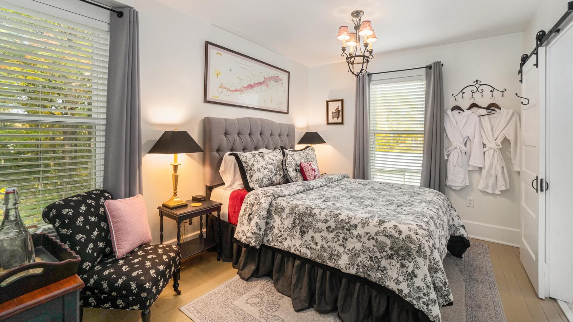 A cozy bedroom featuring a floral patterned bedspread, a black armchair, and bathrobes hanging on the wall.
