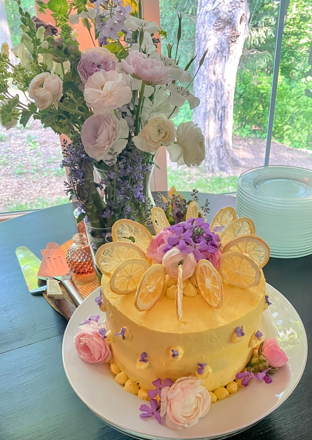 A beautifully decorated lemon cake topped with fruits and flowers, alongside a vase of fresh blooms.