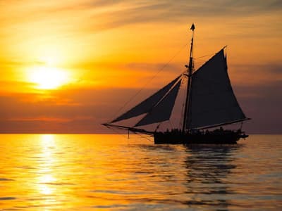 A silhouetted sailing ship glides across a shimmering sea at sunset.