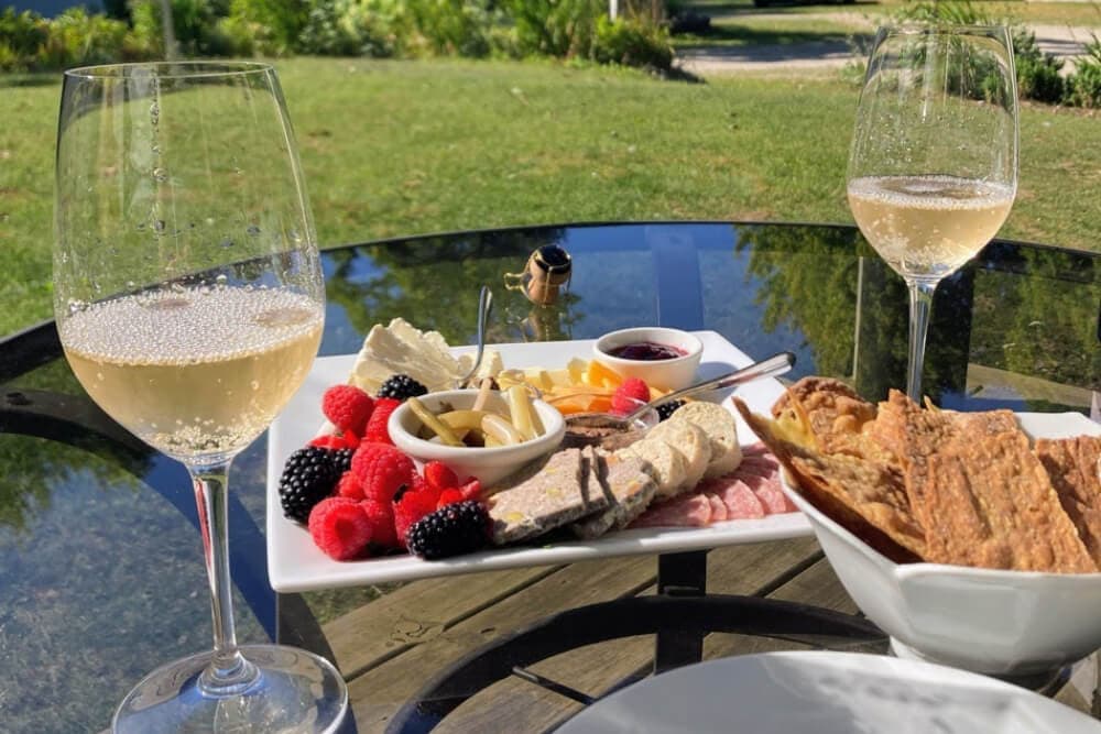 A sunlit outdoor table displays a platter of fruits, cheeses, and snacks alongside two glasses of sparkling wine.