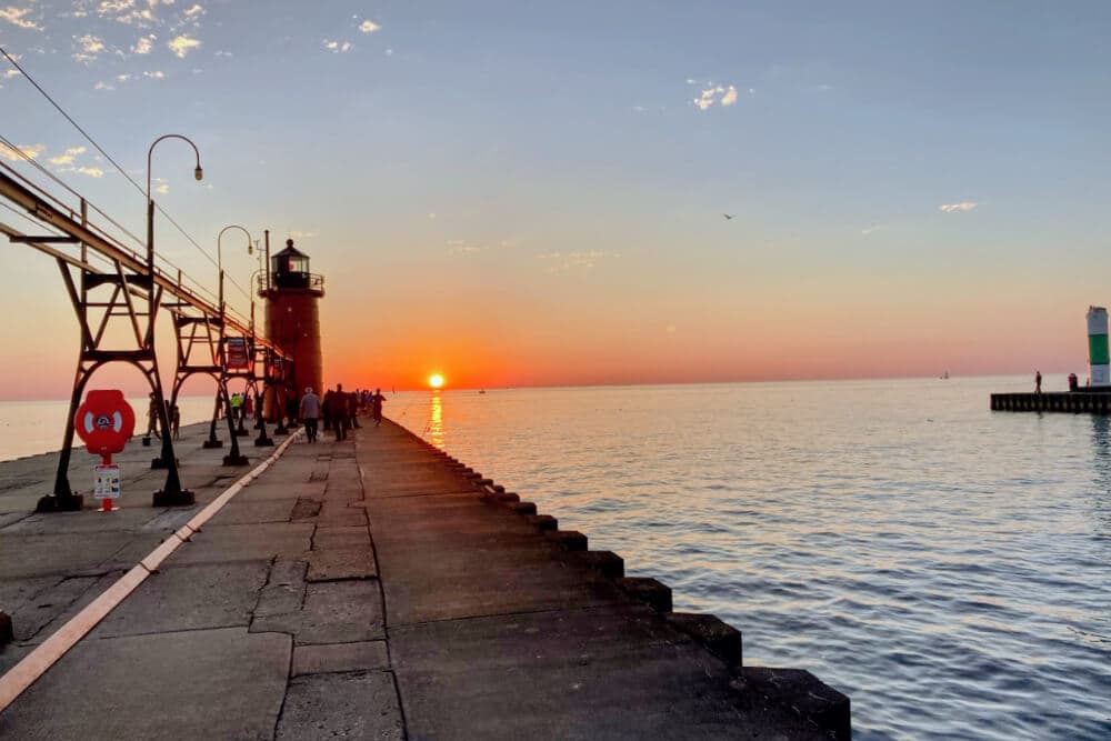 A serene sunset over the water beside a pier and lighthouse, with people strolling along the path.