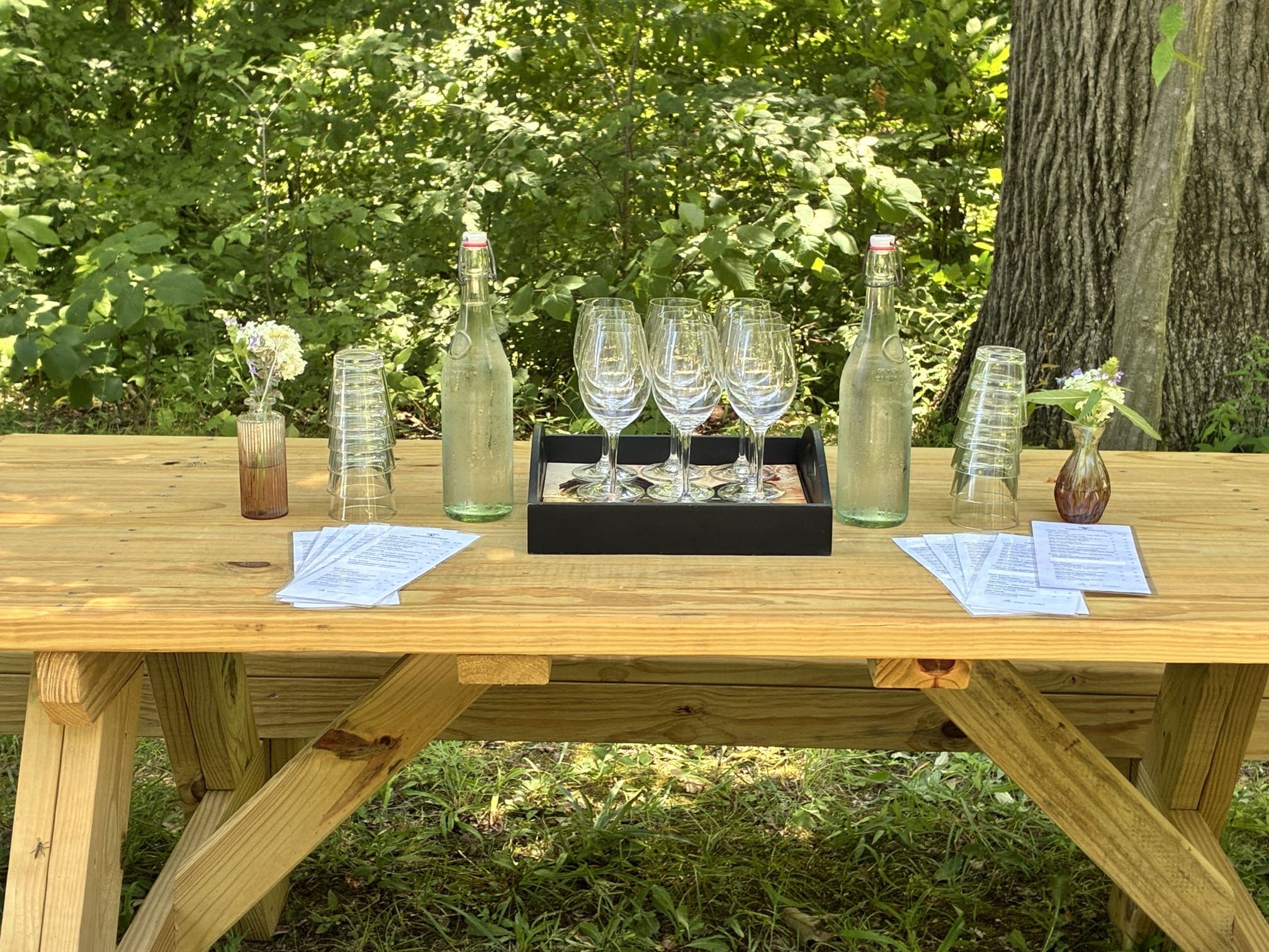 A wooden table set in a lush green area, displaying water bottles, glasses, and menus.