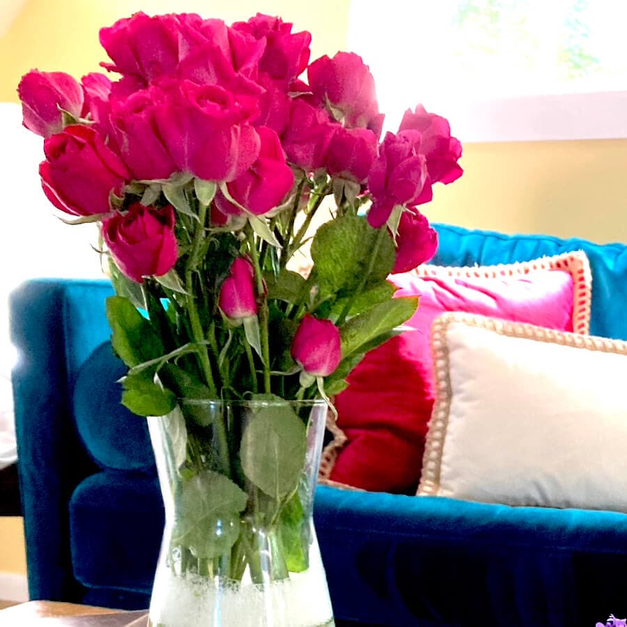 A vibrant bouquet of pink roses in a clear vase sits on a table beside colorful cushions.