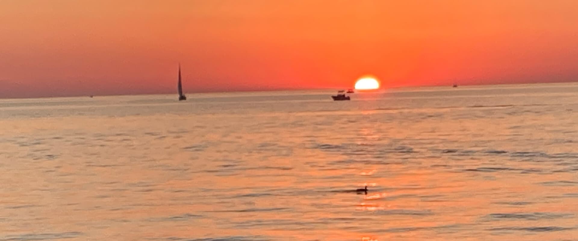 A calm ocean at sunset with a sailboat and a small fishing boat silhouetted against the vibrant orange sky.