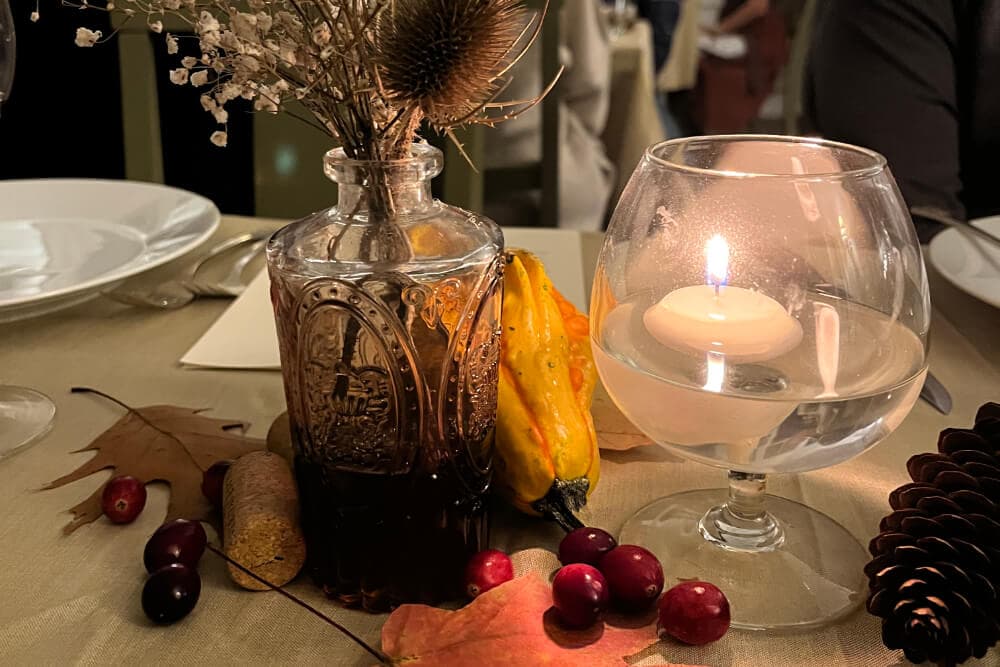 A decorative table setting featuring a glass vase with dried flowers, a candle in a clear holder, gourds, fall leaves, and berries.