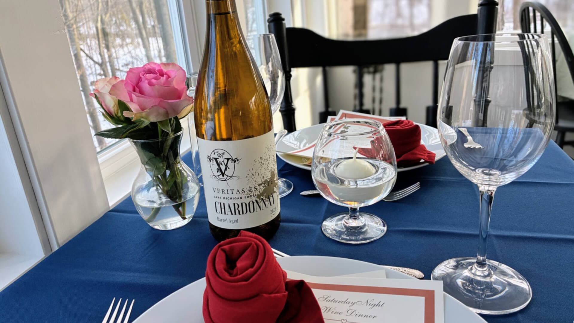 A beautifully set dining table featuring a bottle of Chardonnay, a vase of pink roses, and elegantly folded napkins.