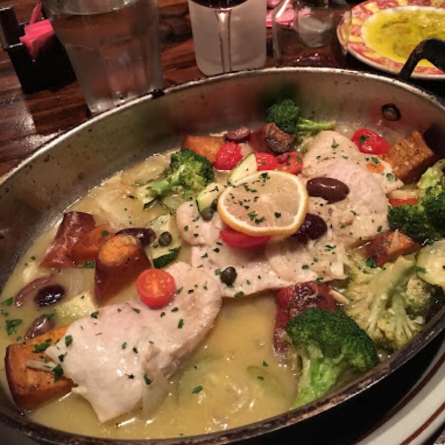 A silver platter filled with cooked fish topped with lemon slices, surrounded by broccoli, tomatoes, and olives in a flavorful sauce.