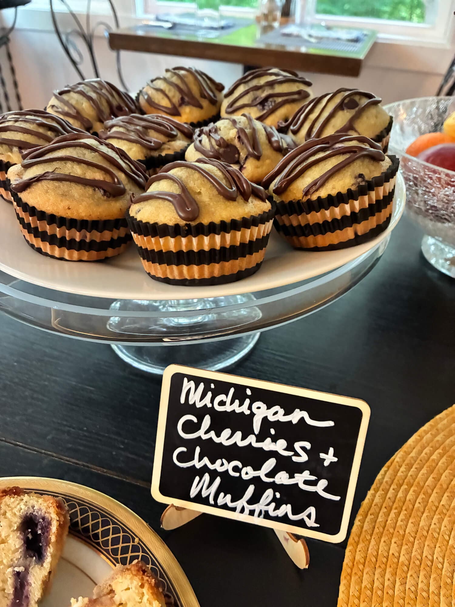 A display of Michigan cherries and chocolate muffins on a glass stand, with a small chalkboard label.