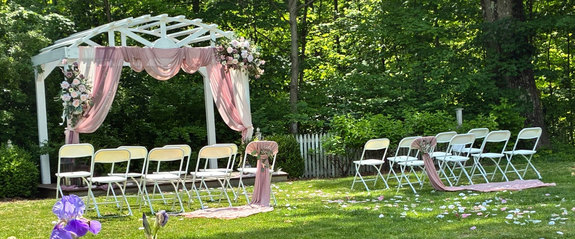 A simple outdoor wedding setup featuring a white pergola adorned with pink drapes and floral arrangements, surrounded by rows of white chairs on a grassy area.