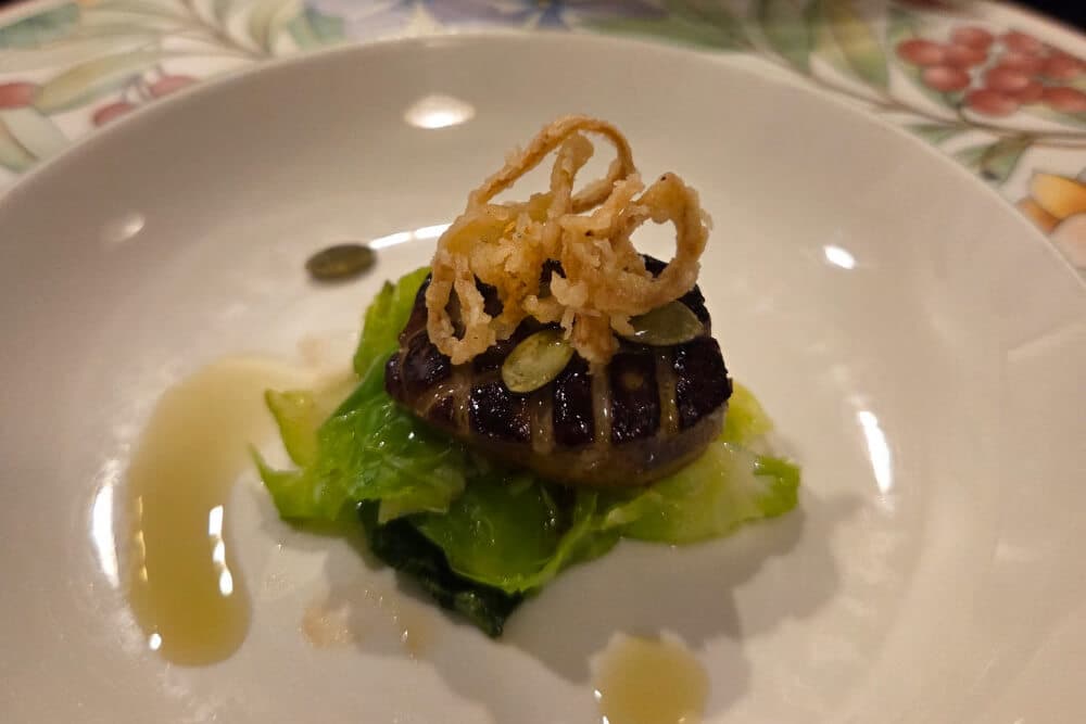 A small plate featuring seared scallops on a bed of sautéed greens, garnished with crispy fried onions and pumpkin seeds.